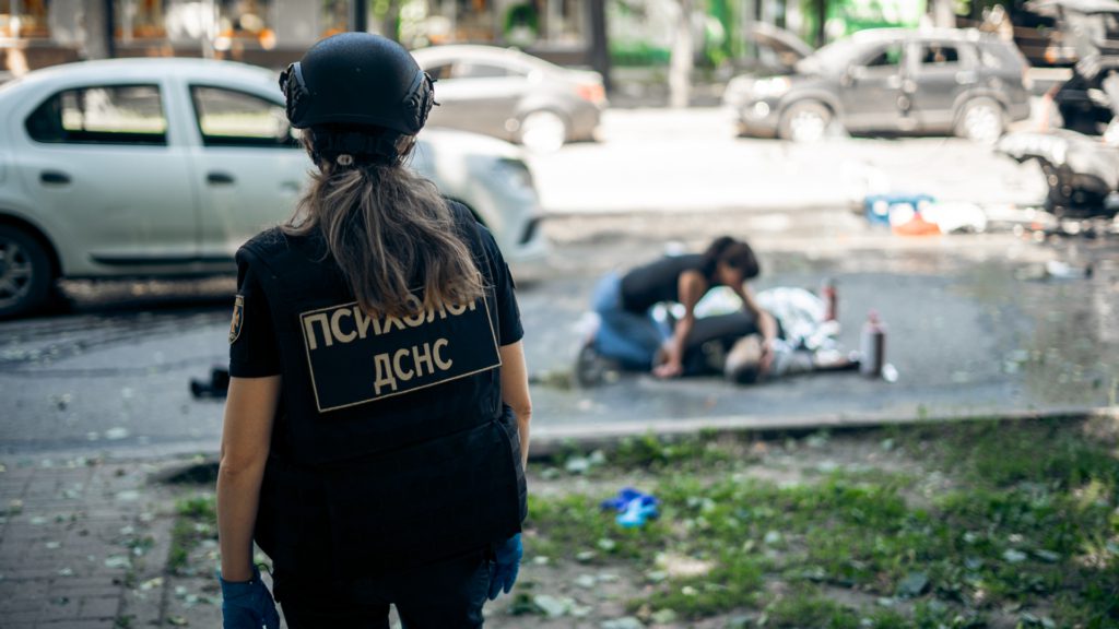 Крупний кадр: психолог ДСНС під час роботи на місці влучання у Сумах. На задньому плані жінка у цивільному одязі схиляється над тілом постраждалого.