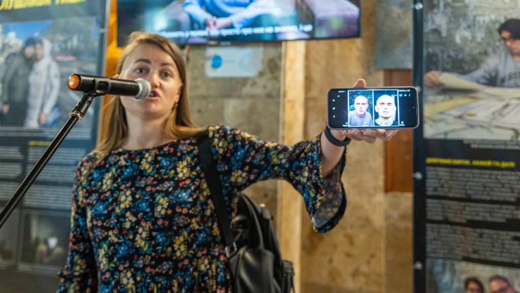 Людина виступає перед відвідувачами виставки, демонструючи на екрані мобільного телефону фото двох осіб, що за словами спікера є близькими людьми та перебувають у російському полоні.
