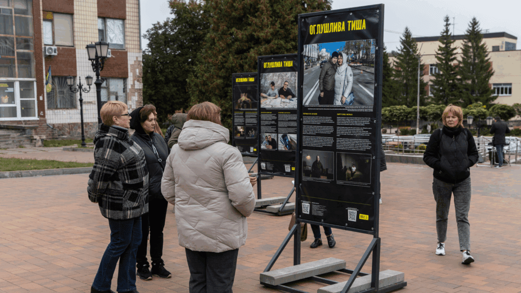 Група людей роздивляється стенди фотовиставки «Оглушлива тиша» на площі, вид збоку