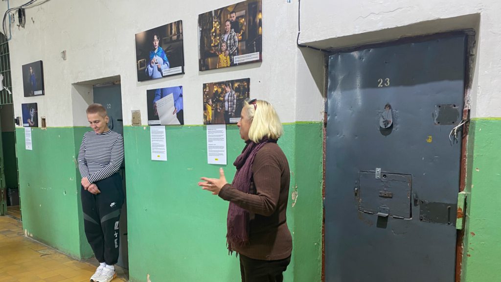 На задньому плані - біло-зелена стіна з масивними тюремними дверима. На стіні розміщені шість фото. На передньому плані двоє осіб, одна з яких звертається до людей поза кадром.