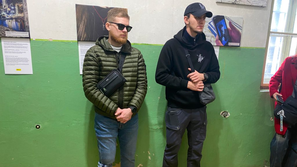 Двоє осіб стоять на тлі стіни, де розміщені фото з експозиції