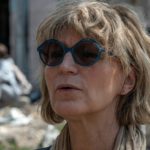 Генеральна секретарка Amnesty International Аньєс Калламар крупним планом. На задньому плані - розмите зображення, у якому проглядається пошкоджений будинок і силует людини, що сидить перед ним.